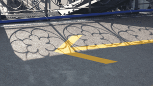 Ausschnitt einer Straße, die untere Hälfte des Weges liegt im Schatten, auf der oberen scheint die Sonne und ein Geländer wirft Schatten, ein gelber Pfeil in der Mitte zeigt nach links