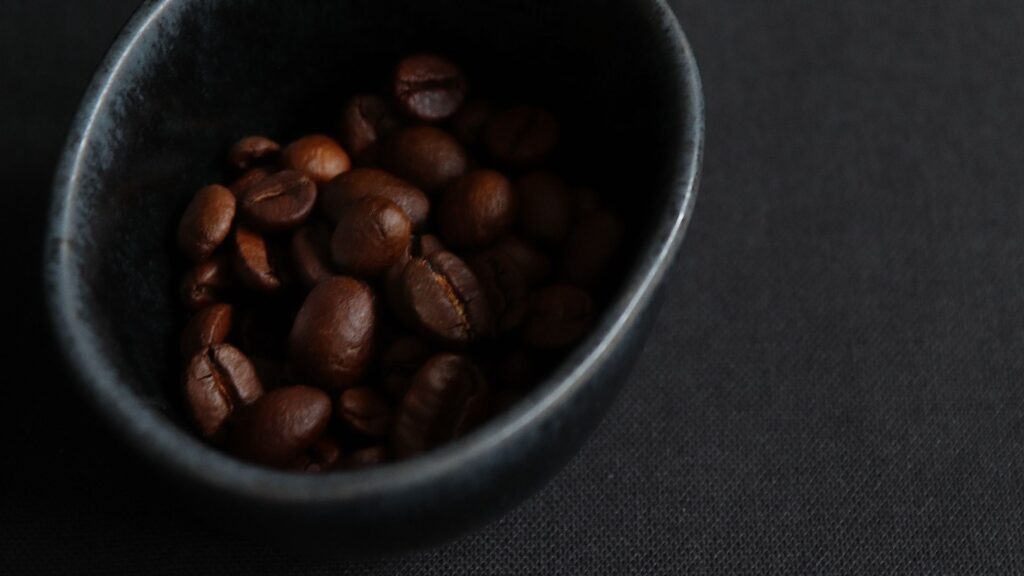 Geröstete Kaffeebohnen in einer anthrazitgrauen Keramik-Schüssel auf dunkelgrauer Tischdecke aus Leinen. Aufnahme im 45° Winkel.